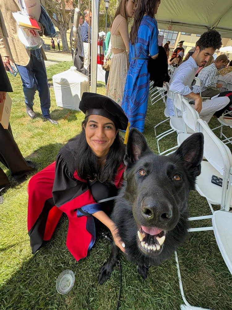 Natalya in a graduation gown, smiling beside her black German Shepherd on the grass